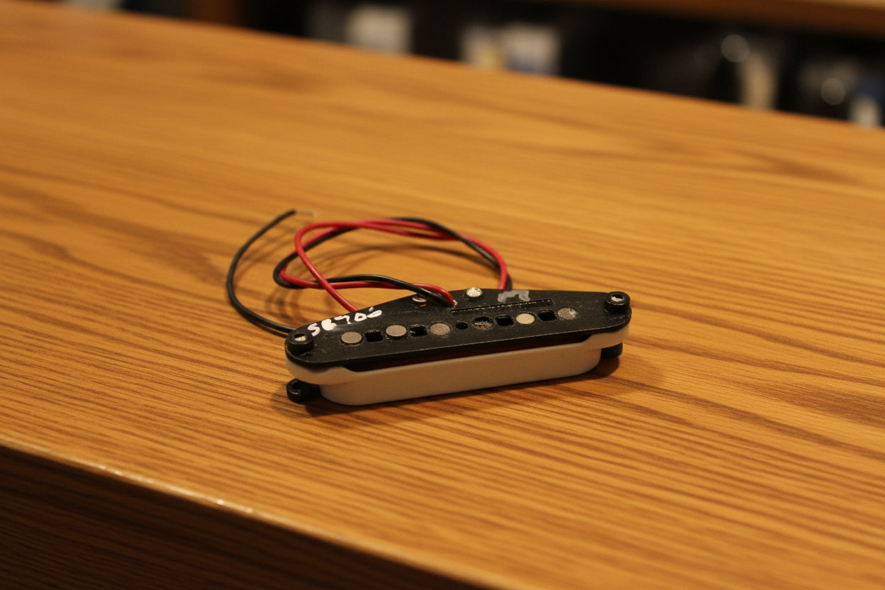 A Fender humbucker guitar pickup with red and black wires, resting on a white fabric background with a Fender logo patch and part of a dark box visible.