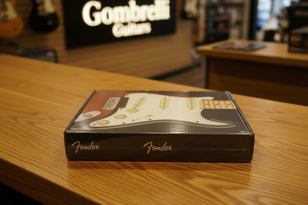 A packaged electric guitar pickguard with a white body, gold hardware, and a maple neck insert, shown inside its clear plastic box on a dark textured surface.