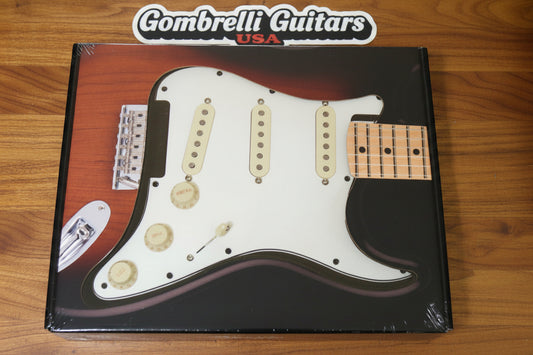 A packaged  vintage 65 Fender electric guitar pickguard with a white body, gold hardware, and a maple neck insert, shown inside its clear plastic box on a dark textured surface.