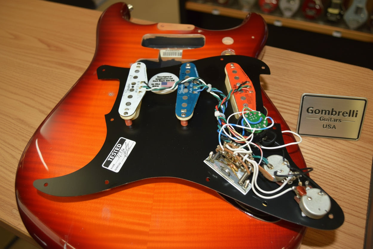 Close-up of a guitar body showing the internal electronics, including three pickups (white, blue, and orange), wiring, and control knobs mounted on a black pickguard over a sunburst orange guitar body.