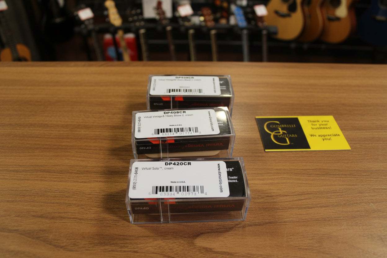 Three small rectangular product boxes stacked vertically on draped champagne satin fabric, with a yellow and black greeting card to the right. The boxes have white labels with black text and barcodes.