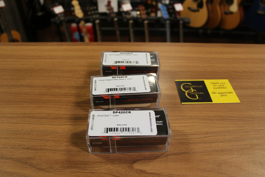 Three small rectangular product boxes stacked vertically on draped champagne satin fabric, with a yellow and black greeting card to the right. The boxes have white labels with black text and barcodes.